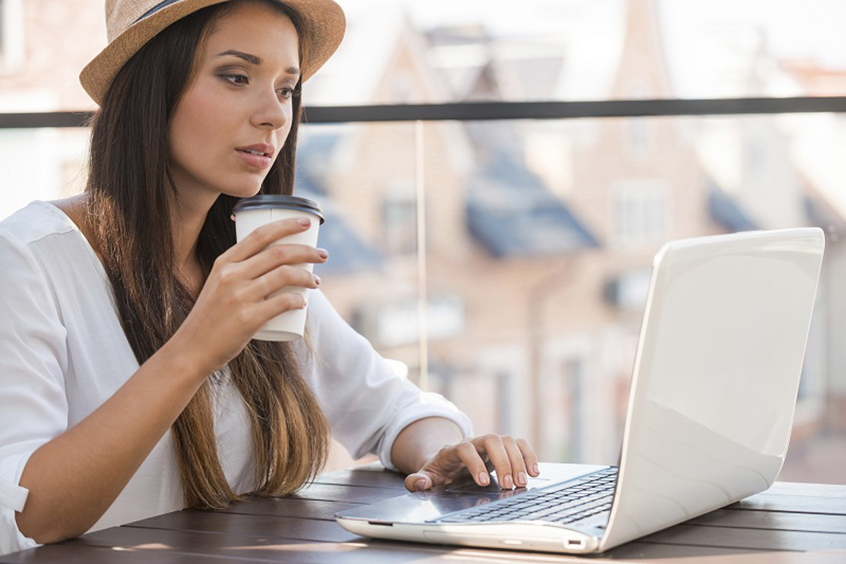 Working outdoors. Beautiful young woman in funky hat working on laptop and smiling while sitting outdoors