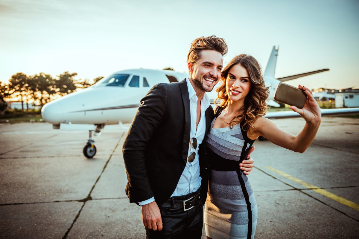 Rich and famous couple taking a selfie for social media while embracing each other in front of a private airplane parked on a tarmac